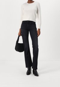 Femme portant un pull blanc, un jean noir à jambes droites, des bottines noires, tenant un sac à main noir texturé sur un fond blanc uni.