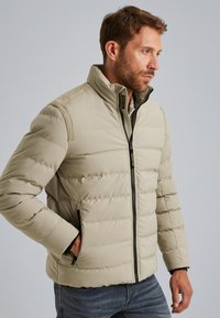 Veste matelassée beige avec un col haut, texture matelassée horizontale, fermeture éclair complète et poches latérales. Matériau doux et isolant.