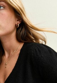 Svart, texturerad tröja med V-ringning. Guldörhängen i form av ringar och lager av guldhalsband med hjärtan och hänge. Ljusbrunt hår som flödar.