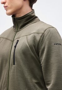 Άνδρας με ελιάς πράσινη ριμπ ύφασμα ζακέτα με φερμουάρ και ψηλό γιακά, με τσέπη στο στήθος με φερμουάρ, κοιτάζοντας αριστερά μπροστά σε απλό φόντο.