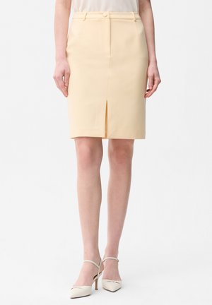 Beige blyantskørtelængde nederdel med frontslids, kombineret med lys top og hvide højhælede sko med ankelstropper.