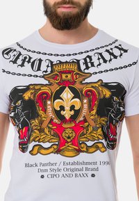 Weißes Baumwoll-T-Shirt mit einem bunten Grafikdesign, das Elemente eines Panthers, eines Lions und einer Krone zeigt, hervorgehoben durch schwarze Kettenmuster.