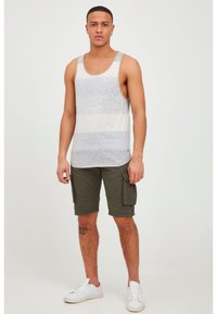 Young man standing wearing sleeveless gray and white striped tank top, dark green cargo shorts, white sneakers, and a tattoo on his left calf.
