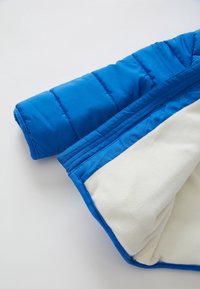 Niebieski rękaw ocieplanej kurtki z widoczną białą podszewką fleece na jasnym tle.