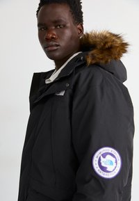 Czarna parka z sztucznym futrem, z zapięciem na zamek i odznaką z napisem "McMurdo Station Antarctica" na prawym rękawie.