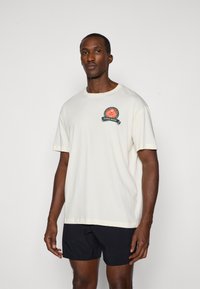 Kremowa koszulka z krótkim rękawem z okrągłym pomarańczowo-zielonym logo na piersi, wykonana z miękkiej bawełny.