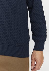 Pulover tricotat bleumarin cu un model texturat, cu manșete și tiv striate, și o etichetă mică pe lateral. Țesătura pare groasă și comodă.