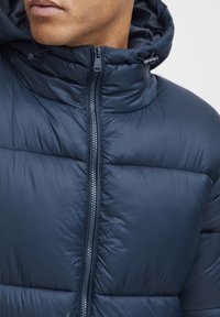 Giacca puffer blu navy con collo alto e chiusura con zip. Realizzata in materiale sintetico, con texture trapuntata e cordino regolabile sul cappuccio.