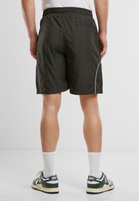Shorts de sport noirs avec une taille élastique, un passepoil blanc sur les côtés et une poche à l'arrière. Porté avec des chaussettes blanches et des baskets vertes.