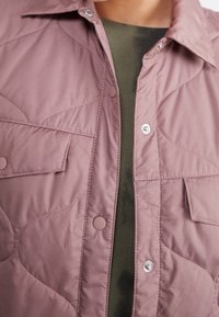 Prošívaná bunda v jemné r�ůžové barvě se sponkovými zapínáním, špičatým límcem a předními kapsami. Nalezena přes texturovaný olivově zelený top.