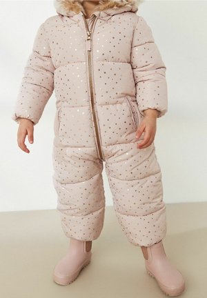 Małe dziecko ubrane w różowy, ocieplany kombinezon zimowy ze złotymi gwiazdkami i kapturem obszytym futerkiem, w różowych butach, stojące wewnątrz pomieszczenia.