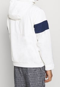 Chaqueta blanca con capucha, presenta una franja azul marino en la parte superior del brazo, textura suave y dobladillo acanalado. Se lleva con pantalones estampados.