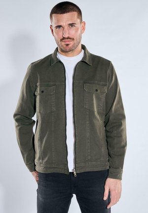 Mann mit kurzen Haaren und Bart, der eine olivgrüne Jacke mit Reißverschluss über einem weißen Hemd und dunklen Hosen trägt, steht vor einem einfachen Hintergrund.