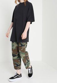 Černé oversized tričko s volným střihem, spojené s kalhotami v zeleno-hnědém maskáčovém vzoru a černými teniskami s bílými podrážkami, stojící na bílém pozadí.