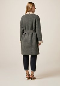 Grahamfarbener Wollmantel mit beigem Kragen, tailliert und mit Gürtel, kombiniert mit dunklen Jeans und Riemchen-Heels.
