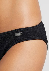 Melna bikini apakšda ar teksturētu ģeometrisku rakstu, ar logotipa birku. Izstrādāta ar zemu jostu un gludu auduma apdari.