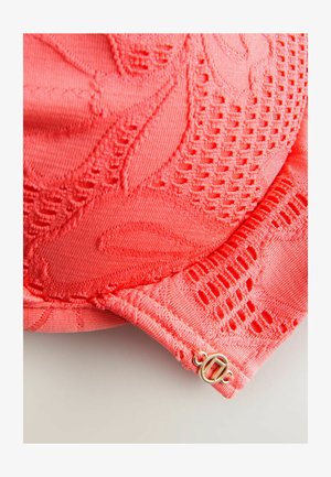 Soutien-gorge en dentelle corail avec des motifs floraux, un tissu texturé et un fermoir doré avec un design circulaire sur le côté.