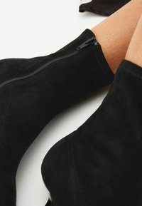 Czarne zamszowe botki za kostkę o gładkiej fakturze, z bocznym zamkiem błyskawicznym i dopasowanym krojem, który przylega do kostki.