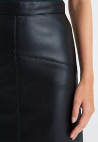 Black leather straight skirt with smooth texture and visible stitching details. Hand resting on the skirt shows its fitted design.