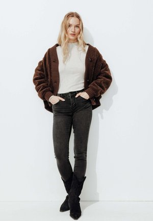 Veste en fourrure marron surdimensionnée avec des poignets côtelés, portée sur un haut ajusté gris clair. Jeans skinny noirs et bottes noires à talons pointus. Fond blanc.