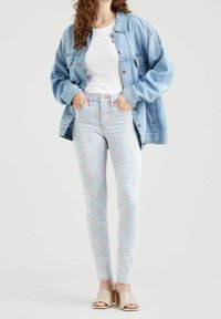 Chaqueta de mezclilla azul claro sobre una camiseta blanca, combinada con unos skinny jeans florales en tonos pastel. La modelo lleva sandalias de tacón beige.