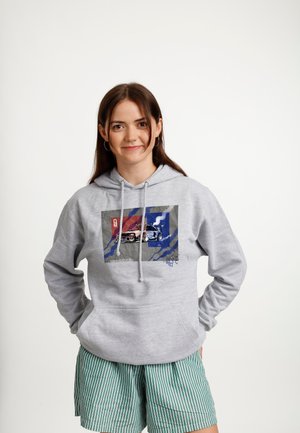 Młoda kobieta w szarej bluzie z kapturem z nadrukiem samochodu i zielonych szortach w paski, stojąca z rękami w kieszeniach na białym tle.
