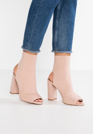 Beżowe sandały na obcasie z otwartymi palcami, z górą wykonaną z dzianiny, wycięciami po bokach oraz masywnym słupkowym obcasem, zestawione z podartymi, krótkimi dżinsami.