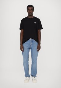 Svart bomull t-skjorte med logo på venstre bryst, kombinert med lyseblå denimbukser og hvite joggesko. Modellen står foran en ensfarget bakgrunn.