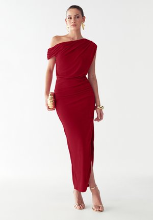 Abito rosso con spalle scoperte, corpino drappeggiato, silhouette aderente, spacco laterale e tessuto morbido; abbinato a accessori dorati e sandali con cinturini.