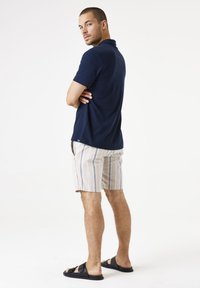 Marineblauwe polo, beige gestreepte shorts en zwarte sandalen. Model staat met gekruiste armen en toont een relaxte pasvorm en casual stijl.