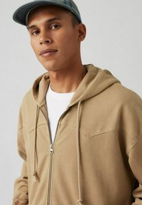 Tan zip-up hoodie met een grote capuchon en trekkoorden, voorzien van voornaden en een zachte textuur, gedragen over een wit T-shirt.