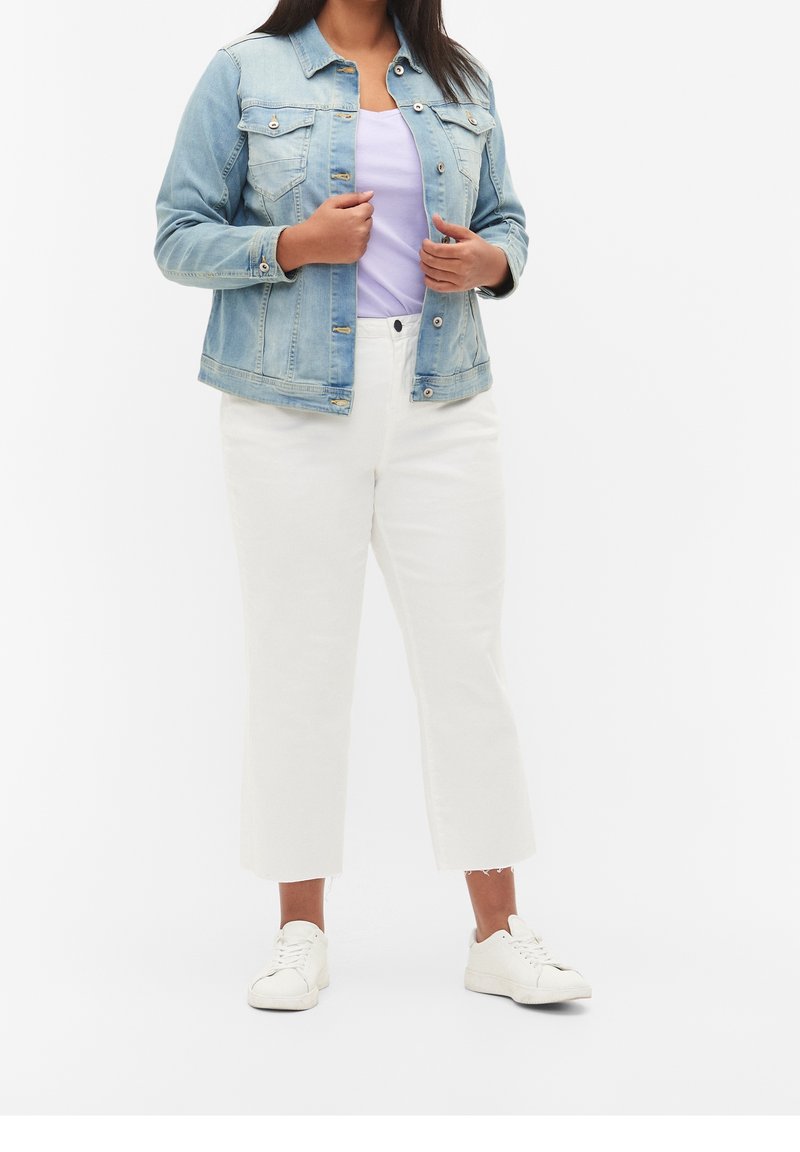 Femme portant une veste en jean bleu clair, une chemise lavande, un pantalon court blanc et des baskets blanches, debout devant un fond clair uni.