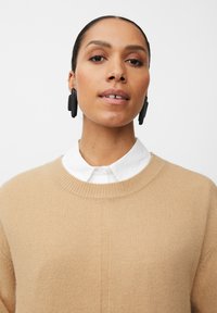 Beige gestrickter Pullover mit Rundhalsausschnitt über einem weißen Hemd mit Kragen. Verfügt über strukturierten Stoff und auffällige schwarze Ohrringe.