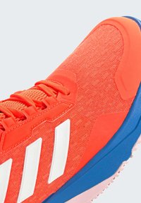 Buty sportowe w jaskrawym pomarańczowym materiale, z białymi paskami. Amortyzowana podeszwa z niebieskimi i różowymi akcentami. Sznurowadła wkomponowane w design.