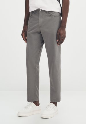 Personne portant un pantalon gris coupe droite, des baskets blanches et une chemise blanche, debout devant un fond clair uni.