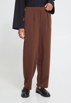 Personne portant un pantalon marron plissé ample, un haut noir à manches longues et des mocassins noirs, debout devant un fond clair et uni.