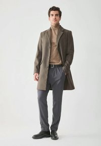 Férfi barna kabátban, világosbarna garbóban, szürke nadrágban és fekete cipőben egy egyszínű világos háttér előtt, kezét a zsebében, előre néz.