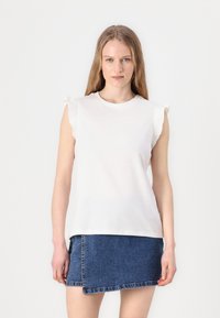Bluză albă fără mâneci, cu umeri volanați, asortată cu o fustă mini din denim cu margini desfăcute. Textură netedă, design casual.