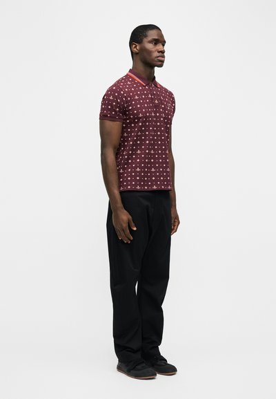 Άνδρας με πουκάμισο πόλο μπορντό με σχέδια, μαύρο παντελόνι και μαύρα παπούτσια, στέκεται με τα χέρια χαλαρά πάνω σε λευκό, απλό φόντο.