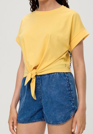 Kvinde iført en gul kortærmet top bundet i taljen og blå denimshorts med buet kant og synlige lommer.