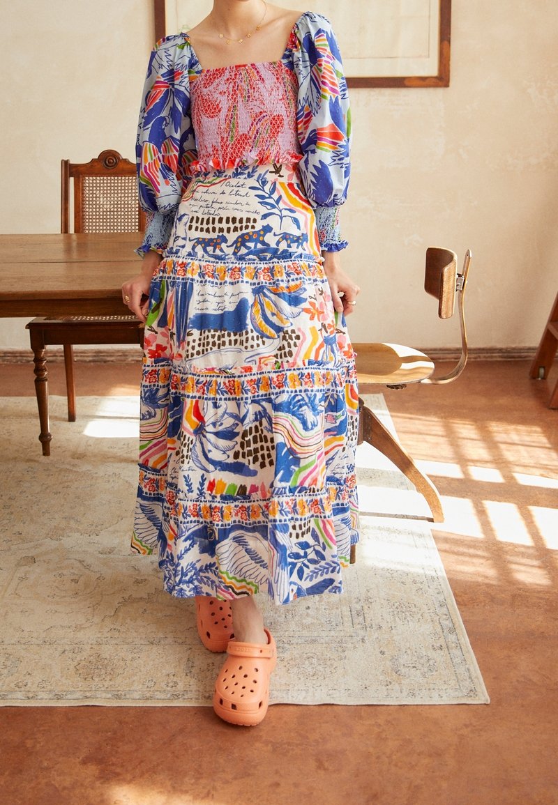 Femme portant une robe colorée à motifs avec des manches bouffantes et des chaussures orange à enfiler, debout sur un tapis près de chaises et d’une table en bois.