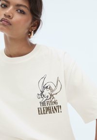 T-shirt en coton crème avec un imprimé graphique noir de Dumbo et le texte "L'ÉLÉPHANT VOLANT !" en typographie en gras.