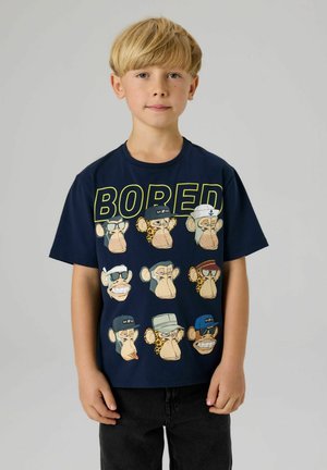 Vaaleahiuksinen poika, joka pukeutunut laivansinisään T-paitaan, jossa on yhdeksän sarjakuvamaista apinan naamaa ja suuri keltainen teksti "BORED" yksinkertaista vaaleaa taustaa vasten.