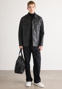 Joven vestido con chaqueta y pantalones de cuero negro, sosteniendo una gran bolsa de cuero negro, de pie sobre un fondo liso con zapatillas blancas.