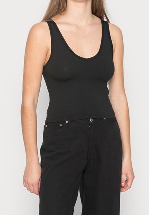 Kobieta ubrana w czarny prążkowany, bezrękawnik oraz czarne spodnie z wysokim stanem, stojąca na tle jasnego, jednolitego tła.