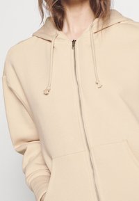 Sweat à capuche beige avec fermeture éclair, fabriqué en tissu doux, doté d'une capuche avec cordon de serrage, de poches avant et d'une fermeture éclair métallique avec un curseur texturé.