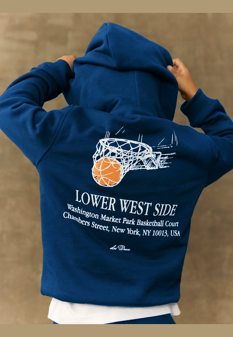 Tums zila kapuce ar basketbola grafiku, kas ietver tīklu un oranžo bumbu, un uz muguras ir teksts "LOWER WEST SIDE".