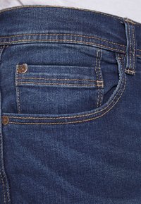 Mörkblå denimjeans med en framficka, med kontrasterande guldstickning och en bronserad nitar detalj nära fickans kant.