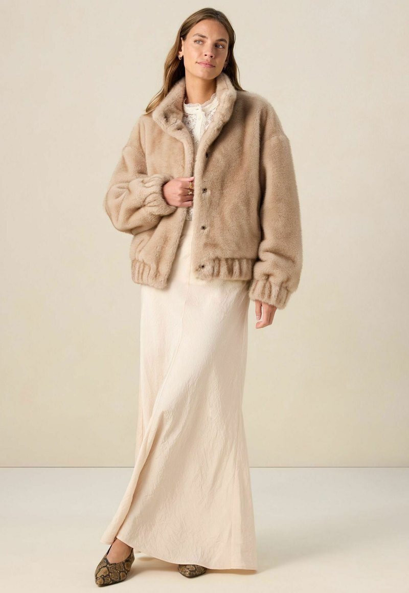 Veste bomber en fausse fourrure beige clair, avec un col, des boutons pression et des poignets côtelés. Portée sur une longue robe crème et des chaussures à motifs.