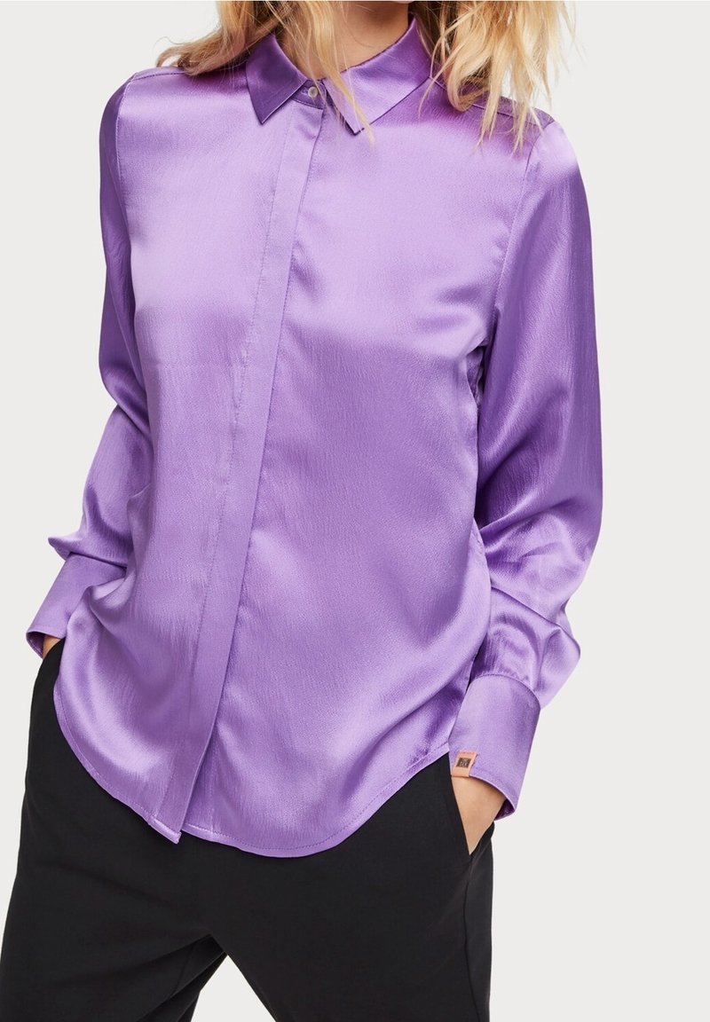 Camicia di raso viola con colletto e maniche lunghe. Presenta una chiusura frontale con bottoni e un orlo arrotondato. Texture liscia e lucida.